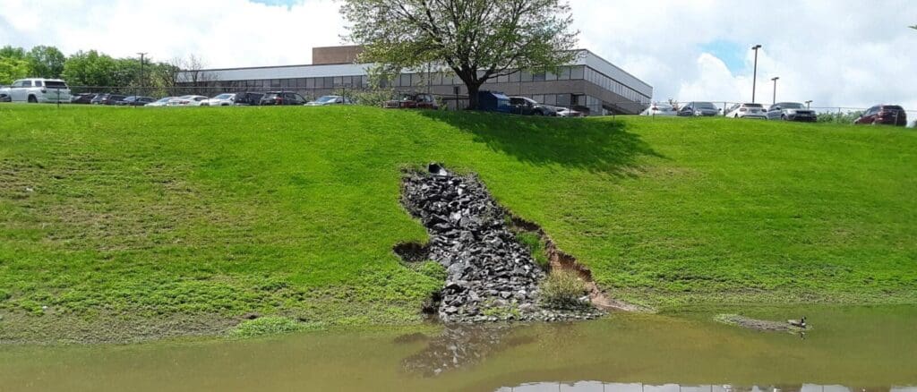 Stormwater detention basin at the foot of grassy hill split by a rock- filled gash below office building and parking lot