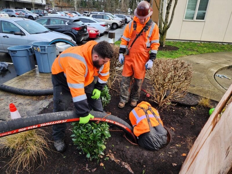 Three men in protective gear insert a large hose into a manhole